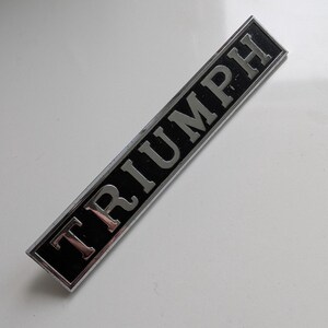 May include: Rectangular black and silver Triumph emblem. The word "TRIUMPH" is in silver capital letters. The emblem has a silver border and is set against a white background.