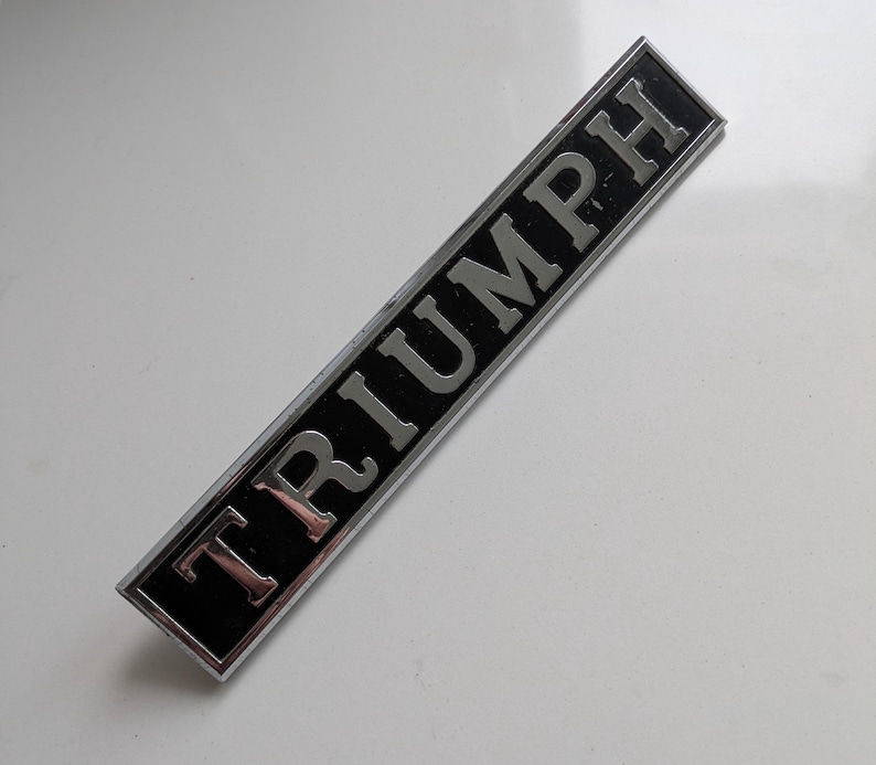 May include: Rectangular black and silver Triumph emblem. The word "TRIUMPH" is in silver capital letters. The emblem has a silver border and is set against a white background.