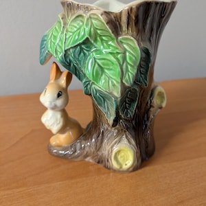 Puede incluir: Un jarrón de cerámica con diseño de tronco de árbol marrón, hojas verdes y un pequeño conejo marrón. El jarrón tiene un interior crema y es adecuado para pequeños arreglos florales. El conejo está sentado en una rama. Aproximadamente 15 cm de alto.