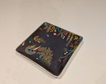 May include: Square decorative dish with a dark background and a gold rim. The dish features a colorful bird and floral design. The edges of the dish are decorated with colorful leaves and flowers.