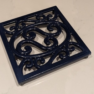 Può includere: Un sottopentola quadrato in metallo blu navy con un design ornato e a spirale. Il sottopentola ha un bordo rialzato e piedini. Questo oggetto decorativo è progettato per proteggere le superfici dalle pentole calde.