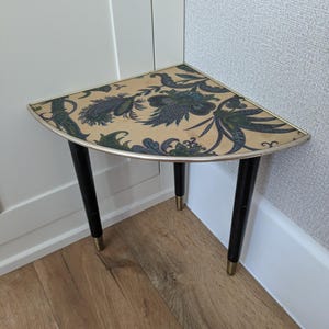 May include: A corner table with a quarter-circle top featuring a beige surface with a dark blue and green floral paisley pattern. The table has three tapered black legs with metallic gold-toned caps at the base and a thin gold-toned metal rim.