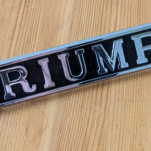 May include: A rectangular Triumph emblem with a black background and silver lettering. The emblem is set against a light brown wooden surface. The word "TRIUMPH" is prominently displayed.