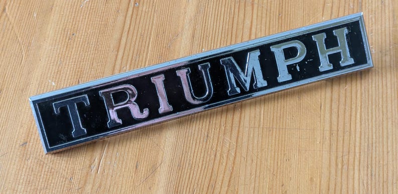 May include: A rectangular Triumph emblem with a black background and silver lettering. The emblem is set against a light brown wooden surface. The word "TRIUMPH" is prominently displayed.