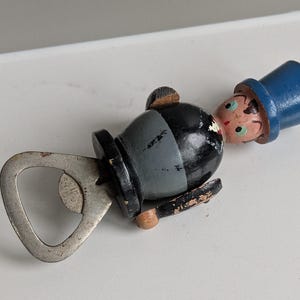 May include: Vintage bottle opener in the shape of a wooden figure with a blue top hat and painted face. The figure has a black body and a silver metal opener.
