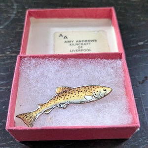 Op de afbeelding: Een visvormige broche met gele en bruine spikkels op een witte vulling in een klein roze kartonnen doosje. Het deksel bevat de tekst AA AMY ANDREWS KILNCRAFT OF LIVERPOOL in zwarte letters op een wit rechthoekig etiket.