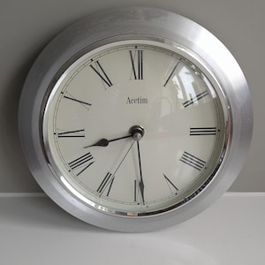 May include: A circular wall clock with a silver-coloured frame and a cream-coloured face. It displays black Roman numerals and black hands. The brand name "Acetim" is printed in black. The clock has a silver bezel.