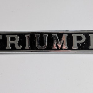 May include: A rectangular Triumph emblem with a black background and silver lettering. The word "TRIUMPH" is displayed prominently. The emblem has a silver border and is set against a white background.