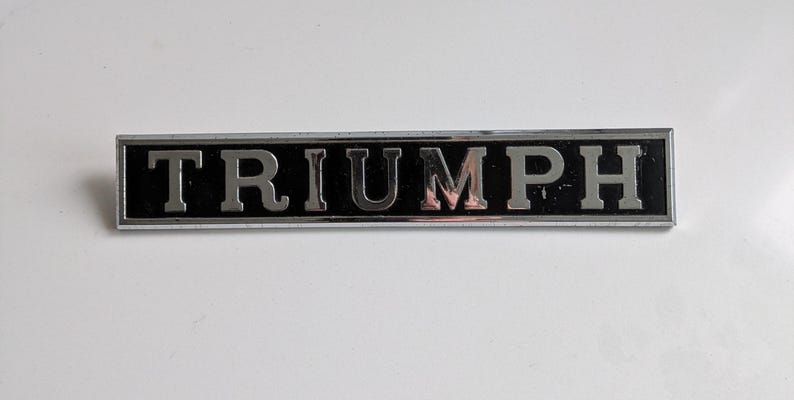 May include: A rectangular Triumph emblem with a black background and silver lettering. The word "TRIUMPH" is displayed prominently. The emblem has a silver border and is set against a white background.