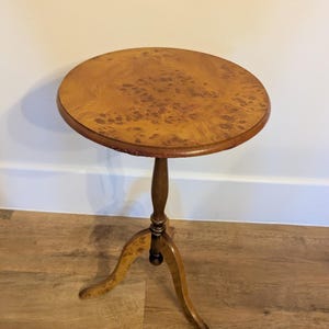 Puede incluir: Una pequeña mesa auxiliar redonda de madera con base de tres patas. La tapa es de color marrón claro con un diseño estampado. Las patas y el soporte central son de color marrón oscuro. La mesa mide unos 61 cm de altura.