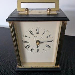 May include: A vintage Timemaster quartz clock with a rectangular shape. The clock has a gold-colored handle and trim, a black base, and a cream-colored face with Roman numerals. The clock's face reads "Timemaster".