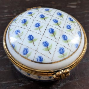 Könnte beinhalten: Eine runde Schmuckdose aus Porzellan mit weißem Untergrund und einem sich wiederholenden Muster aus kleinen blauen Blütenknospen in einem goldfarbenen Gitter. Der Deckel hat einen goldfarbenen Metallrand und einen blumenförmigen Verschluss.
