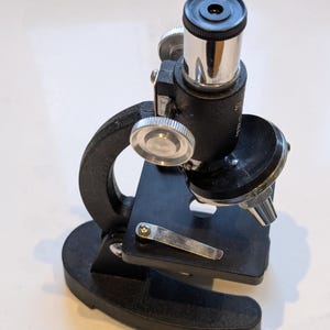 May include: A vintage black microscope with a silver eyepiece and adjustment knobs. The base is rectangular, and the arm is curved. The microscope is designed for scientific study and observation.
