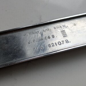 May include: A rectangular, silver-colored metal plate with engraved text. The text includes "J. FRAY LTD. BHAM", "J.F. 4149.", and "P.N. 621078.". The plate has a slightly worn appearance, suggesting age or use. A small, circular attachment is visible on the left side.