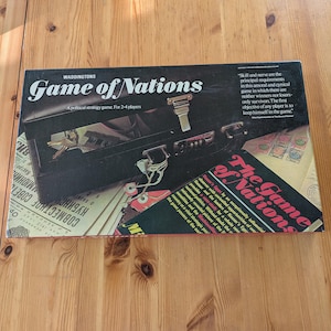 以下が含まれることがあります： Waddingtonsの「Game of Nations」というヴィンテージボードゲームの箱。 黒い箱には白い文字とブリーフケースの画像があります。 このゲームは2〜4人用の政治戦略ゲームで、テキストも含まれています。