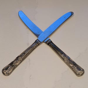 Op de afbeelding: Twee zilveren botermessen gekruist, de messen reflecteren licht. De handvatten hebben een sierlijk, bloemmotief. De messen staan tegen een neutrale achtergrond, wat wijst op een formele eetgelegenheid. De messen zijn gebogen en lijken scherp.