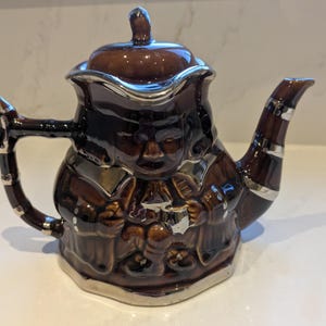 Könnte beinhalten: Eine braune Keramik-Teekanne mit silberner Verzierung und Deckel. Die Teekanne hat ein einzigartiges Design mit einem Gesicht und Figuren auf dem Körper. Der Griff und der Ausguss sind ebenfalls braun mit silbernen Akzenten. Ein dekorativer Küchenartikel.