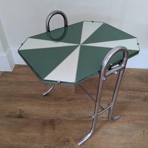 May include: A side table featuring a rectangular glass top with a geometric sunburst pattern in alternating dark green and white segments. The table is supported by a polished chrome metal frame with curved legs and two integrated handle-like loops.