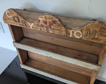 Puede incluir: Estantería de especias de madera con dos niveles. La parte superior tiene las palabras "COSY HOME" y una ilustración de vaca. La estantería es de madera marrón claro con detalles blancos.