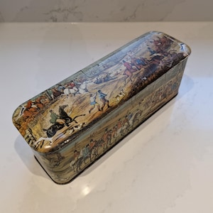 Puede incluir: Caja de lata rectangular con una ilustración detallada de una escena de caza. La caja presenta una tapa y laterales curvados, con una estética vintage. La obra de arte representa personas a caballo, perros y figuras en un campo, con una paleta de colores apagados.