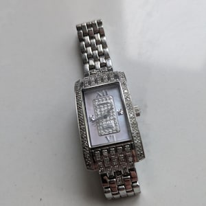 Puede incluir: Reloj de pulsera rectangular plateado con esfera blanca y detalles de diamantes. El reloj tiene una correa plateada y una esfera rectangular con números romanos. Un artículo de lujo.