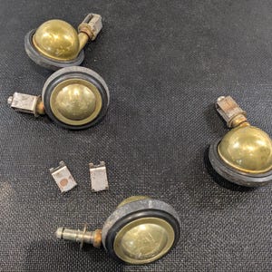 May include: A set of antique brass castors with black rubber tyres and mounting hardware. The castors have a ball shape and are designed for furniture. The brass has a vintage patina, and the rubber tyres show signs of wear.