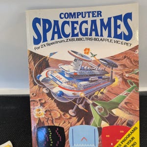May include: A retro book cover for "COMPUTER SPACEGAMES" featuring a sci-fi illustration of spaceships and a planet. The text includes compatibility for ZX Spectrum, ZX81, BBC, TRS-80, APPLE, VIC & PET. Includes game program graphics.