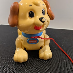 Può includere: Un giocattolo a forma di cucciolo Fisher-Price giallo con orecchie marroni, un collare blu e un cordino rosso. Il giocattolo ha occhi neri e una lingua rossa che sporge. Il logo Fisher-Price è visibile sul corpo del giocattolo.