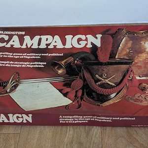 Könnte beinhalten: Eine Vintage-Brettspielbox mit dem Titel "CAMPAIGN" in Rot und Weiß. Die Box zeigt Illustrationen militärischer Gegenstände wie ein Schwert, einen Helm und eine Rüstung. Das Spiel wird als militärisches und politisches Strategiespiel beschrieben.