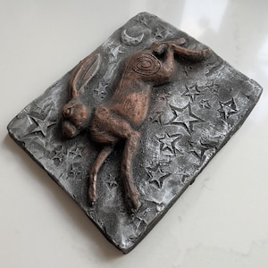 Könnte beinhalten: Bronzefarbene Hasen-Skulptur auf strukturiertem, grauem Hintergrund mit Sternen und einem Mond. Der Hase springt, mit zurückgelegten Ohren und ausgestreckten Beinen. Die Skulptur ist auf einem rechteckigen Sockel montiert.