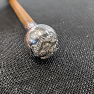 May include: A silver-colored walking stick handle with a wooden shaft. The handle is round with a detailed emblem that reads "The Own Royal West Kent Regiment". The handle is silver and the shaft is brown.