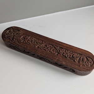 Può includere: Una scatola rettangolare in legno marrone scuro con intricate incisioni floreali. La scatola ha estremità arrotondate e un coperchio incernierato. Le incisioni dettagliate corrono lungo la parte superiore e i lati.