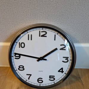 May include: A round wall clock with a white face and black numerals. The clock has a dark metal frame and black hands. The word "quartz" is printed on the face. The clock is approximately 12 inches in diameter.