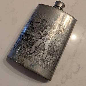 Könnte beinhalten: Eine silberfarbene Flasche mit Schraubverschluss. Die Flasche zeigt eine geätzte Illustration einer Person beim Angeln, die eine Angelrute und ein Netz hält. Die Flasche hat eine rechteckige Form und ein leicht abgenutztes Aussehen.