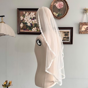 May include: A sheer, white bridal veil with a delicate lace trim is draped over a mannequin. The veil is layered, creating a soft, flowing effect. The mannequin is a neutral beige color, and the background features framed artwork.