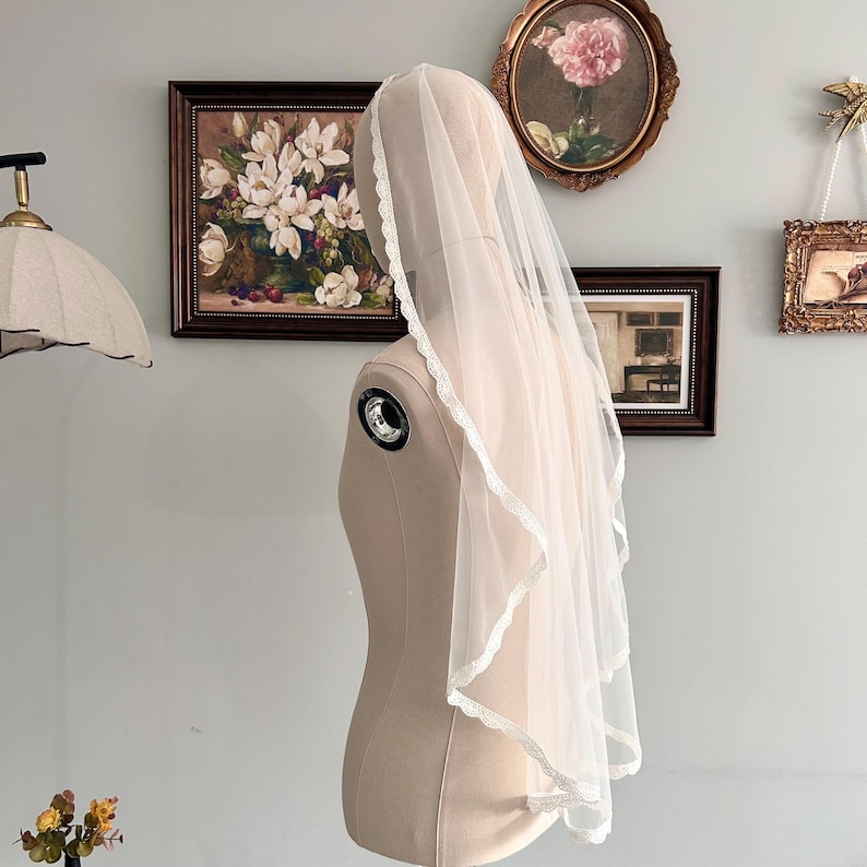 May include: A sheer, white bridal veil with a delicate lace trim is draped over a mannequin. The veil is layered, creating a soft, flowing effect. The mannequin is a neutral beige color, and the background features framed artwork.