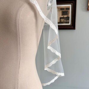 May include: A sheer white bridal veil with delicate lace trim. The veil is draped over a mannequin, showcasing its flowing design and intricate details. The lace trim adds a touch of elegance to the bridal accessory.