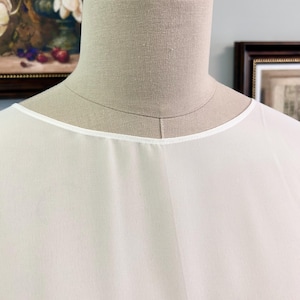 Puede incluir: Primer plano de una blusa blanca y transparente con cuello redondo. La tela parece ligera y ligeramente texturizada. El escote est&aacute; ribeteado con una fina banda blanca. La blusa se muestra en un maniqu&iacute;.