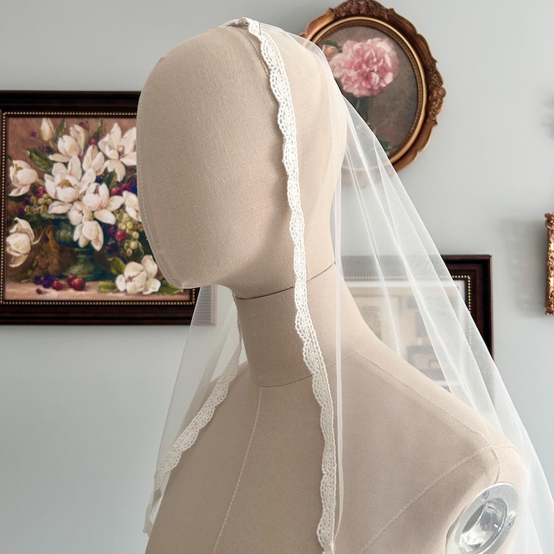 May include: A sheer white bridal veil with a delicate lace trim. The veil is draped over a mannequin head, showcasing its flowing design. The lace trim adds a touch of elegance to the simple, yet beautiful, wedding accessory.