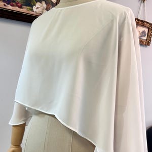Puede incluir: Una blusa tipo capa blanca y transparente con cuello redondo y dise&ntilde;o en capas. La tela cae sobre los hombros y los brazos, creando una silueta fluida. La blusa se exhibe en un maniqu&iacute;.