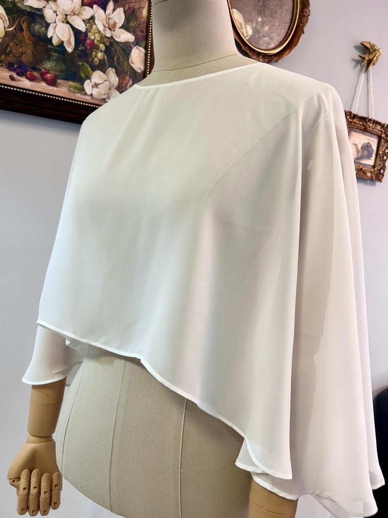 Puede incluir: Una blusa tipo capa blanca y transparente con cuello redondo y dise&ntilde;o en capas. La tela cae sobre los hombros y los brazos, creando una silueta fluida. La blusa se exhibe en un maniqu&iacute;.
