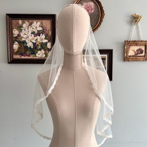 May include: A sheer, white bridal veil with a delicate lace trim. The veil is displayed on a mannequin, showcasing its soft, flowing fabric and elegant design. The lace trim adds a touch of sophistication to this classic wedding accessory.