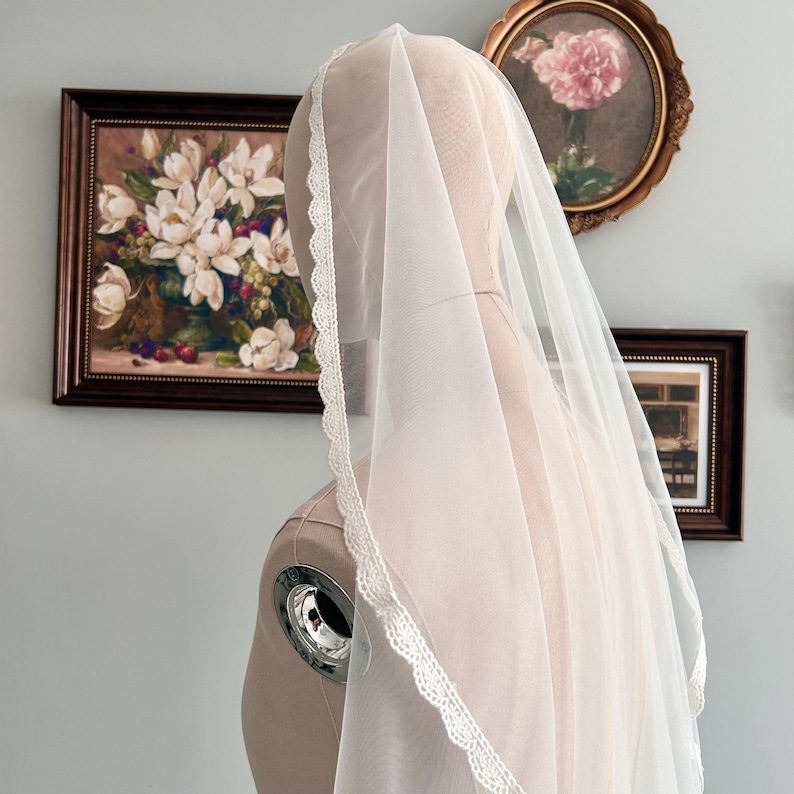May include: A sheer white bridal veil with a delicate lace trim, draped over a mannequin. The veil is lightweight and airy, with a soft, flowing appearance. The background includes framed floral artwork and a light blue wall.