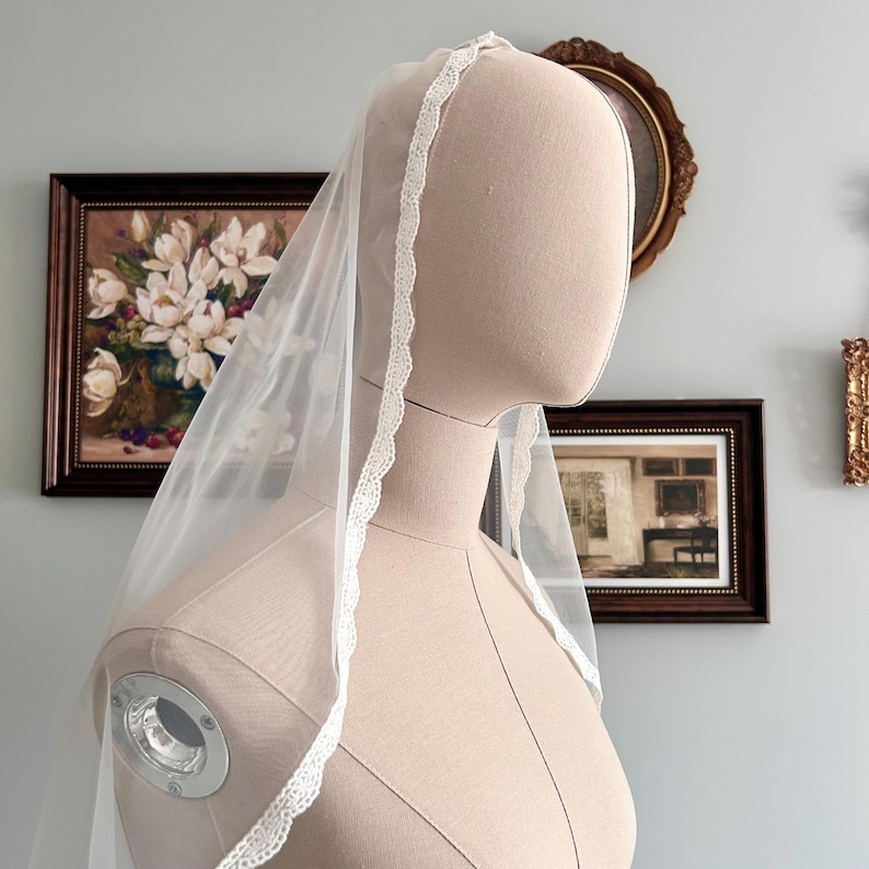 May include: A sheer white bridal veil with delicate lace trim. The veil is draped over a mannequin, showcasing its flowing design. The lace trim adds a touch of elegance to the veil's edge. The veil is a classic accessory for weddings.