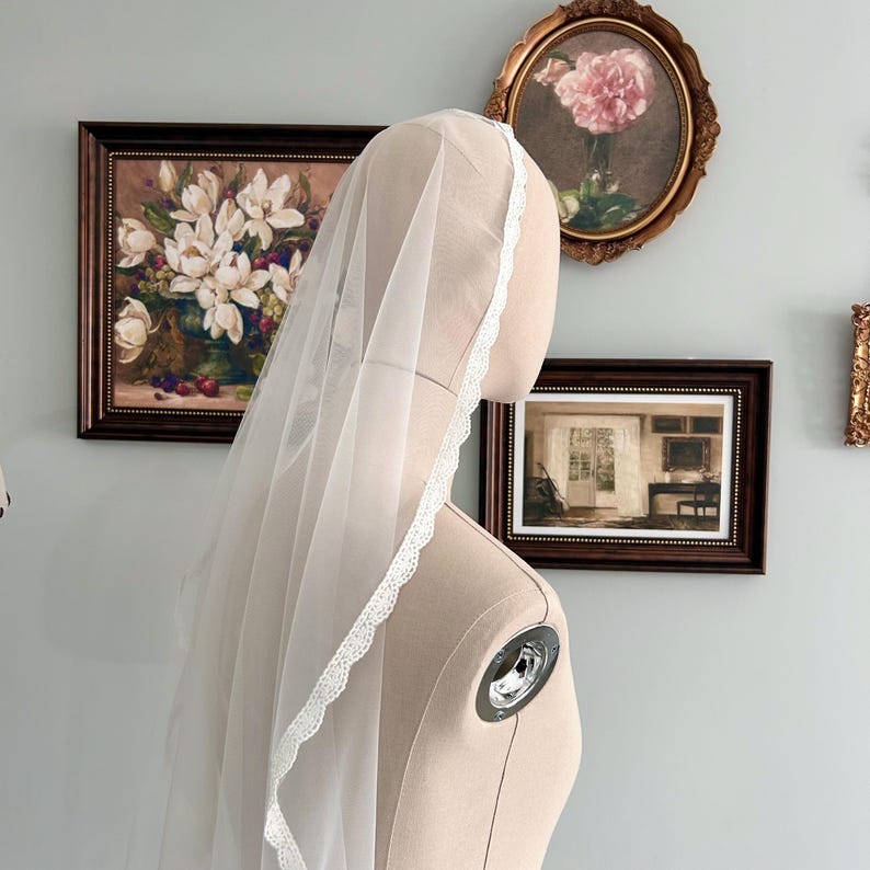 May include: A sheer white bridal veil with delicate lace trim is draped over a mannequin head. The veil is long and flowing, with a soft, ethereal appearance. The background features framed artwork and a light blue wall.