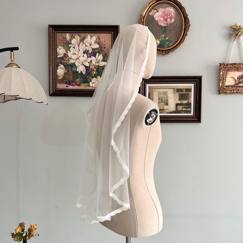 May include: A sheer white bridal veil with a delicate lace trim. The veil is draped over a mannequin, showcasing its length and design. The lace trim adds a touch of elegance to the simple, flowing fabric. The veil is a classic accessory for weddings.