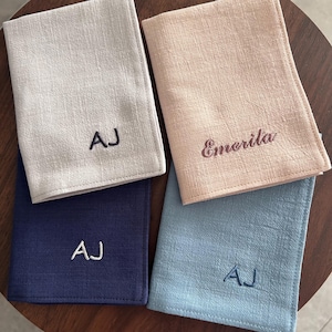 May include: Four personalized linen napkins in shades of gray, navy, peach, and blue. Each napkin is embroidered with initials or a name in a contrasting color. The napkins are folded and arranged on a dark wooden surface.
