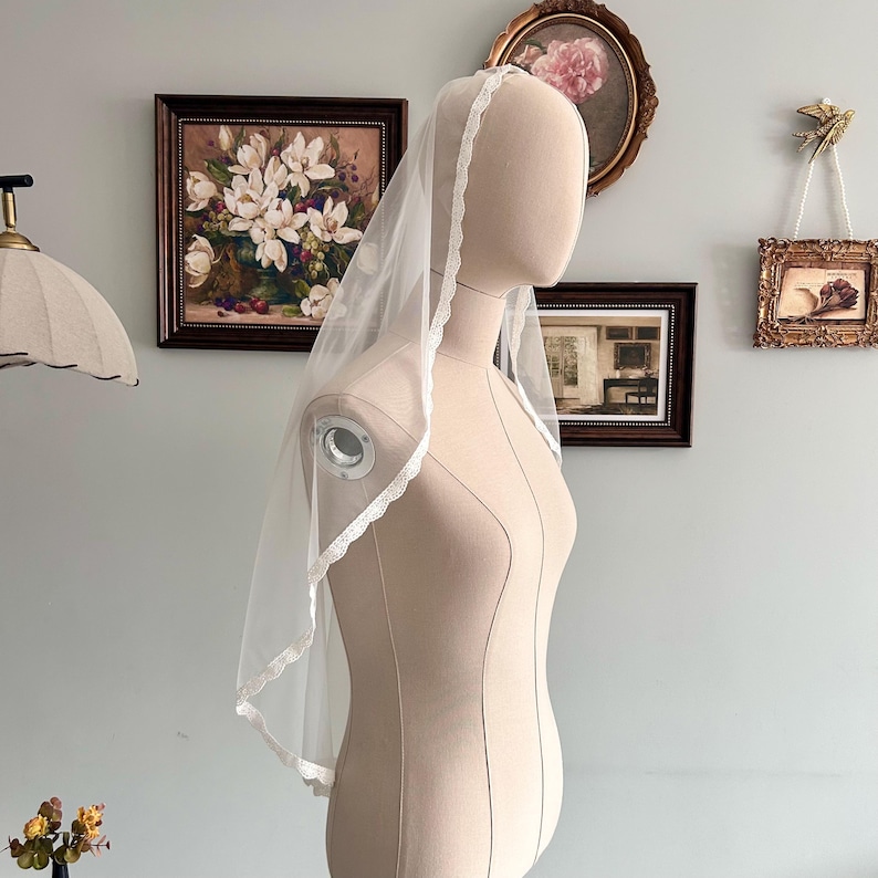 May include: A sheer white bridal veil with a delicate lace trim. The veil is draped over a mannequin, showcasing its shape and design. The background includes framed artwork and a lamp.