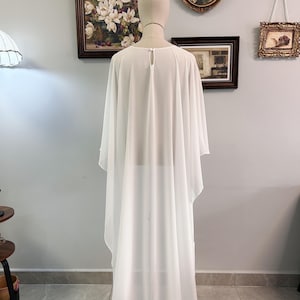 Puede incluir: Un vestido capa blanco y transparente con mangas anchas. El vestido se exhibe en un maniqu&iacute;, mostrando su elegante ca&iacute;da y tejido ligero. El vestido tiene un detalle de ojo de cerradura en la parte posterior del cuello.