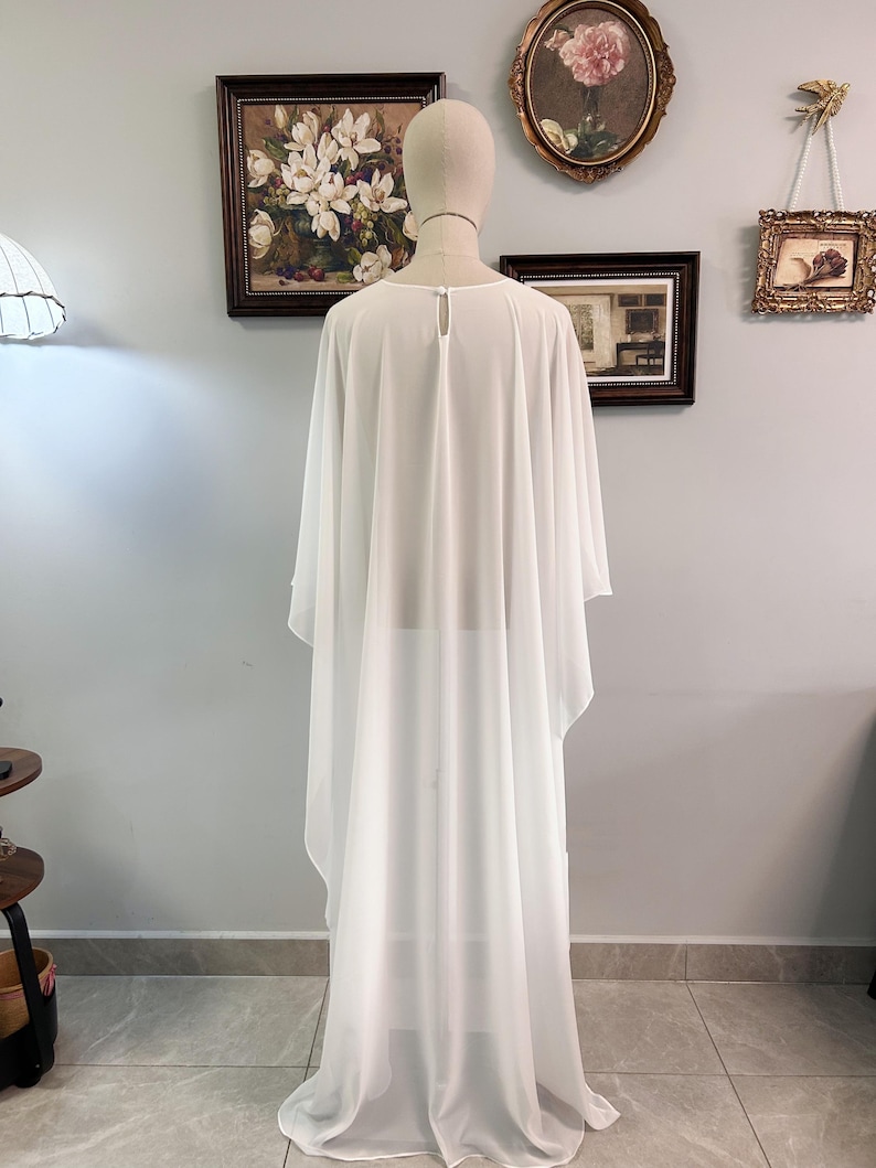 Puede incluir: Un vestido capa blanco y transparente con mangas anchas. El vestido se exhibe en un maniqu&iacute;, mostrando su elegante ca&iacute;da y tejido ligero. El vestido tiene un detalle de ojo de cerradura en la parte posterior del cuello.